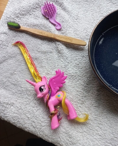 a photo depicting the process: a bowl with water on the top left, toothbrush on the right and cadance with one part of her main twisted up and wrapped around her leg and the other part wet, and layed down straight like a ribbon